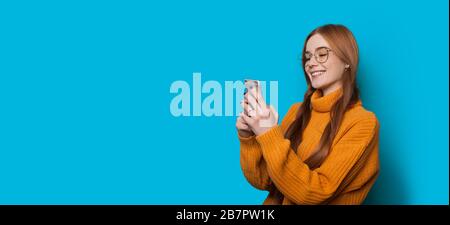 Ginger caucasian girl with freckles and eyeglasses chatting with somebody using a mobile while posing on a blue wall with blank space Stock Photo