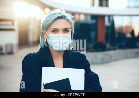Beautiful business woman protecting herself from viruses by wearing special mask while posing outside with her computer and notebook Stock Photo