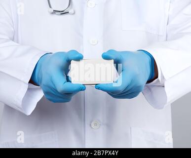 adult doctor therapist is dressed in a white robe uniform and blue sterile gloves is standing and holding a stack of empty white paper business cards, Stock Photo