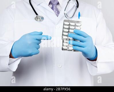 adult doctor therapist is dressed in a white uniform coat and blue sterile gloves is standing and holding a stack of pills in blister packs, white bac Stock Photo