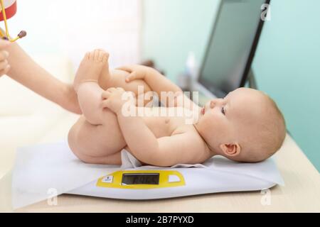 Pediatrician specialist taking measurement infant child weight during screening examination. Cute caucasian baby lying on electronic scales doctors