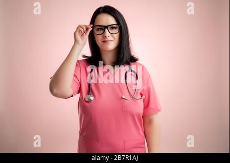 Portrait of beautiful woman doctor with glasses and stethoscope wearing pink scrubs, posing on a pink isolated backround. Stock Photo