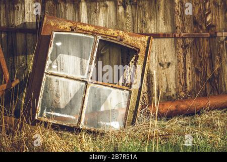 Old rusty and neglected window frame with only three of four window glass remaining. Rusty surroundings, grass is growing around. Nature's trying to r Stock Photo