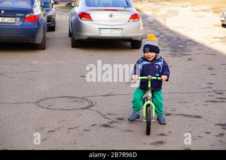 POZNAN, POLAND - Mar 08, 2020: Small two years old boy sitting on a balance bike on asphalt with parked cars. Stock Photo