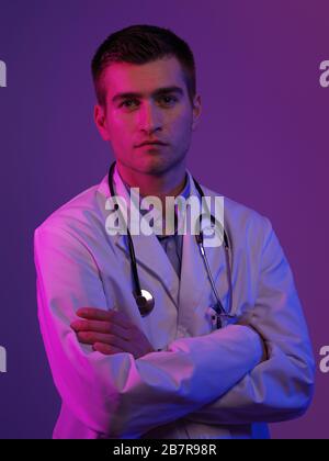Portrait of hero in white coat.  Cheerful smiling young doctor with stethoscope in medical hospital standing against blue and pink background. Coronav Stock Photo