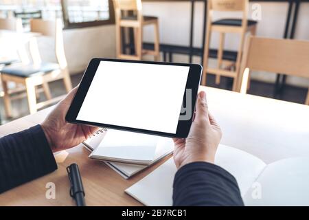 Mockup image of hands holding and using black tablet pc with blank white desktop screen while working on notebooks in office Stock Photo