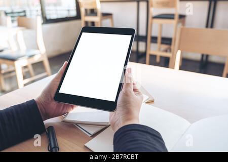 Mockup image of hands holding and using black tablet pc with blank white desktop screen while working on notebooks in office Stock Photo