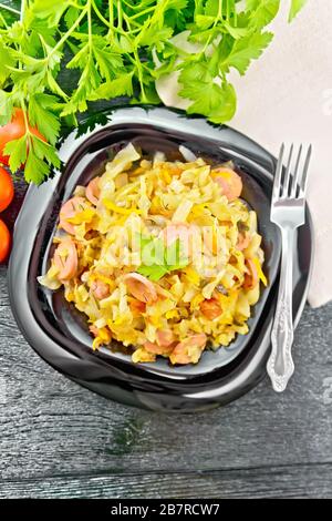 Cabbage stew with sausages in a black plate, napkin, tomatoes, parsley and fork on a dark wooden background from above Stock Photo