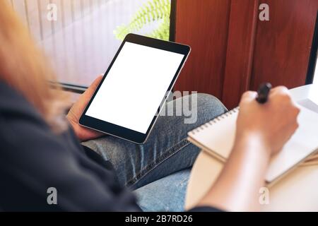 Mockup image of a woman holding and using black tablet pc with blank white desktop screen while writing on notebooks in office Stock Photo