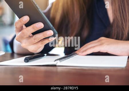 Closeup image of a woman holding and using smart phone with notebook and pen on wooden table Stock Photo