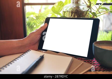 Mockup image of hand holding and using black tablet pc with blank white desktop screen with notebooks on the table Stock Photo