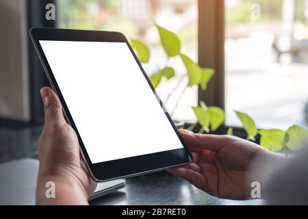 Mockup image of hands holding black tablet pc with blank white desktop screen with green leaves on wooden table Stock Photo