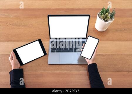 Top view mockup image of a hands holding mobile phone , black tablet and laptop with blank white screen on wooden table in office Stock Photo