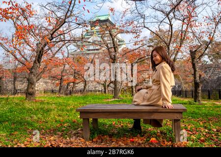 A beautiful asian woman enjoyed sitting on a wooden branch in the autumn garden with Osaka castle in background Stock Photo