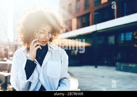 Stunning caucasian woman with curly hair and eyeglasses is wearing a white shirt and having a phone discussion Stock Photo