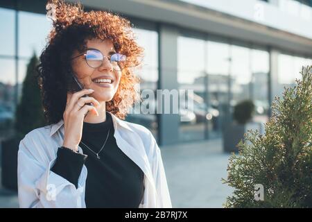 Lovely caucasian freelancer with curly hair is having a phone discussion while walking outside Stock Photo