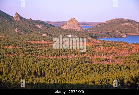 Famous Okzhetpes rock rises above the spruce and birch forest on the shore of Burabay lake at sunset in autumn season Stock Photo