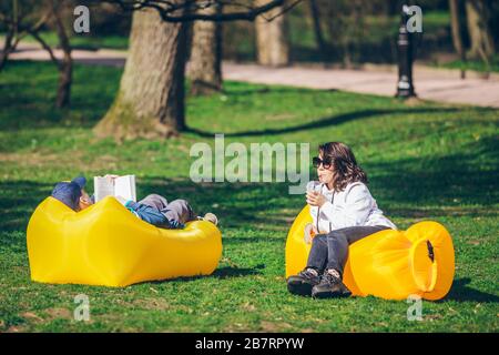 couple laying on yellow inflatable mattress in city park. reading book. drinking smoothies Stock Photo