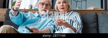 Panoramic shot of senior man pointing with hand by shocked wife while watching tv on couch Stock Photo