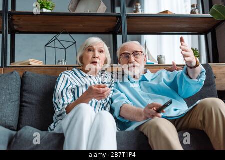 Low angle view of shocked woman watching tv by husband with remote controller on sofa Stock Photo