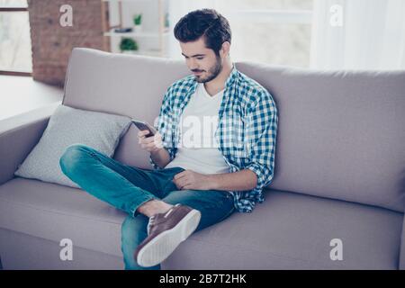 Handsome young smiling man in casual clothes sitting on sofa and using mobilephone for chatting with his girlfriend Stock Photo