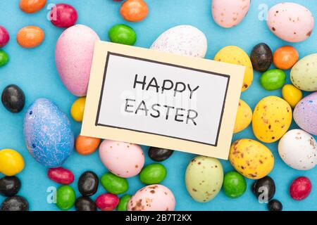 Top view of card with happy easter lettering on colorful chicken and quail eggs, candies on blue background Stock Photo