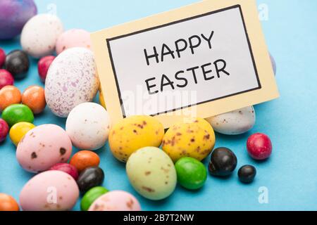 Close up of card with happy easter lettering on bright candies, colorful chicken and quail eggs on blue background
