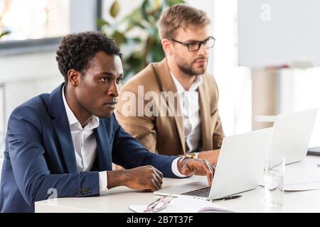 two multiethnic businessmen using laptops at table in office Stock Photo