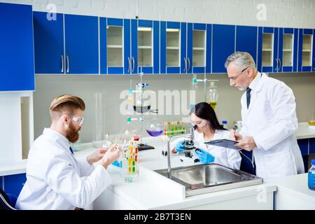 Teamwork concept. Professor is checking and controlling the group work of two lab interns. All three are focused on experiment and wearing labcoats Stock Photo