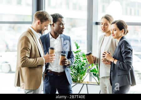 four multiethnic colleagues holding disposable cups of coffee and smiling in office Stock Photo