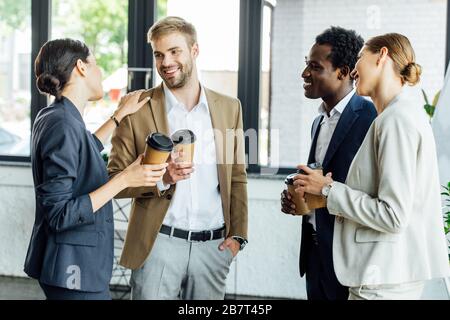 four multiethnic colleagues holding disposable cups of coffee and smiling in office Stock Photo