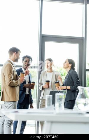 four multiethnic colleagues holding disposable cups of coffee and talking in office Stock Photo