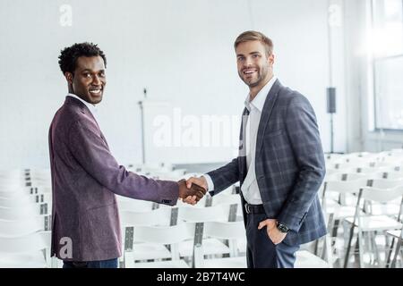 two multiethnic colleagues in formal wear shaking hands in conference hall Stock Photo