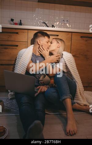 Newly wedded couple kissing each other during a sunny morning while sitting on the floor in the kitchen and using a laptop Stock Photo