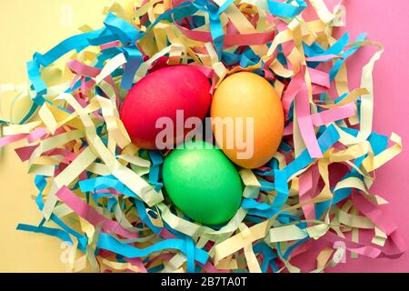 Three dyed Easter eggs in a colorful nest on mixed pink and yellow background. Top view. Symbol of main Christian feast celebration. Stock Photo