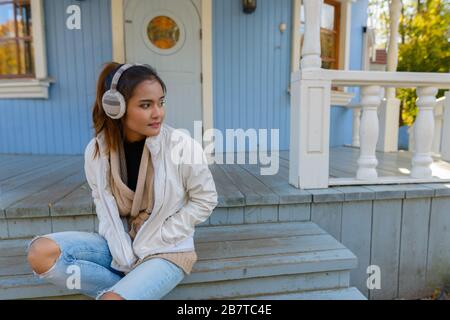 Young beautiful Asian woman thinking and sitting on stairs of old blue house Stock Photo