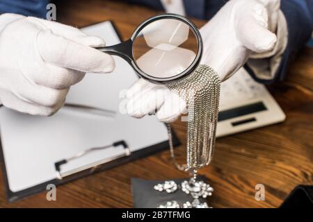 Cropped view of jewelry appraiser holding necklace and magnifying glass near calculator, clipboard and earrings on table Stock Photo