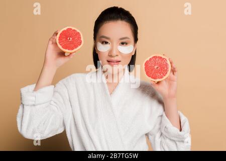 beautiful asian woman in bathrobe with eye patches on face holding grapefruit halves on beige background Stock Photo