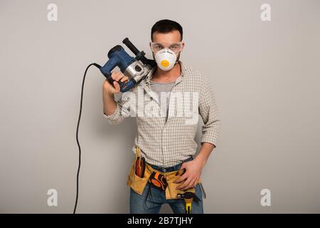 confident manual worker in safety mask holding electric drill on grey Stock Photo