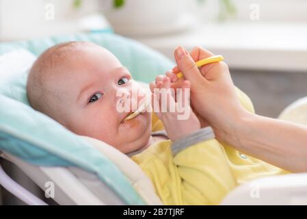 Mom feeds baby vegetable puree in a high chair. The mother puts a spoon of mashed in the child's mouth. The infant eats food. Stock Photo