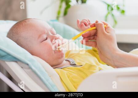 Mom feeds baby vegetable puree in a high chair. The mother puts a spoon of mashed in the child's mouth. The infant eats food. Stock Photo