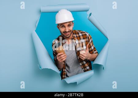 happy repairman in helmet holding coffee to go and showing thumb up in torn paper, isolated on blue Stock Photo