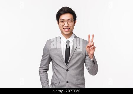 Portrait of successful determined and confident young asian man feeling he can get the deal for enterprise, standing in gray suit, showing peace sign Stock Photo