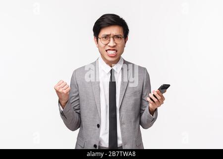 Waist-up portrait of displeased, bothered and annoyed asian businessman in grey suit, clenching teeth and fists, frowning aggressive, grimacing Stock Photo