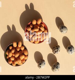 Five quail eggs and two bowls with nuts with shadows like rabbits on the light yellow background. Trendy conceptual minimalistic photo for Easter Holi Stock Photo