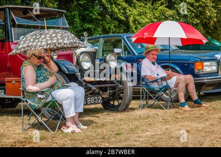 Elderly couple sitting under parasols, (the gent looks a bit grumpy) near a classic car. Enjoying a day at Henley-on-Thames. Traditional Boat Rally. Stock Photo