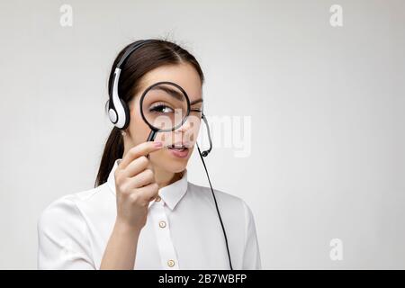 search concept. Beautiful woman consultant of call center in headphones holding magnifying glass on gray background. female customer support operator with headset. I will help find everything Stock Photo