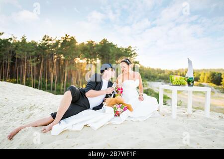 Side view of charming young couple bride and groom clink glasses of champagne while sitting at table. Fruits and bouquet of flowers on background of r Stock Photo