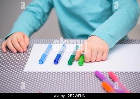 Close-up of five year old boy playing and lining up color pens on a white paper placed on a table with white dots. The boy is holding a green pen. Stock Photo