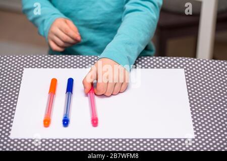 Close-up of five year old boy playing and lining up color pens on a white paper placed on a table with white dots. The boy is holding a pink pen. Stock Photo